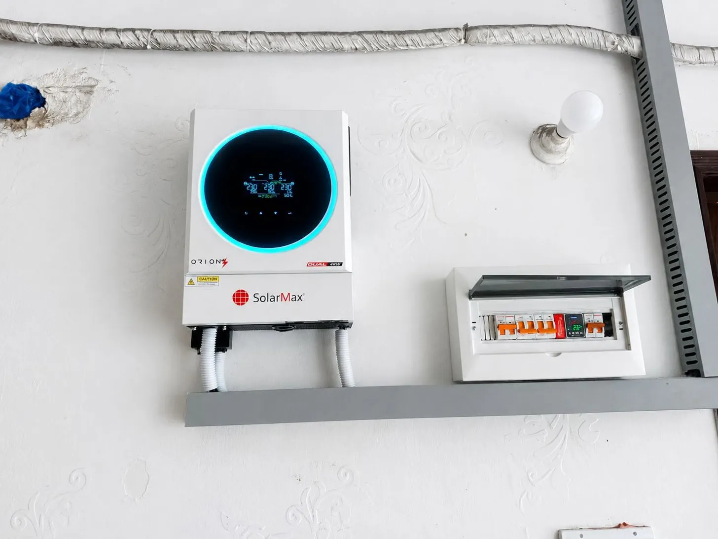SolarMax ORION DUAL 4kW hybrid inverter wall-mounted beside a distribution board with MCBs and a digital voltage protector